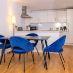 Bright dining area with round table, blue accent chairs, and fully fitted white kitchen at The Sycamore Suite, Enterprise Place, Woking