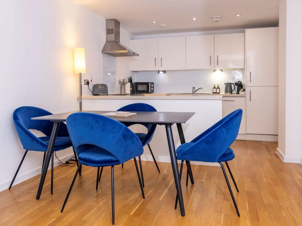 Bright dining area with round table, blue accent chairs, and fully fitted white kitchen at The Sycamore Suite, Enterprise Place, Woking