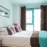 Bright double bedroom with pink cushions, artwork, and modern window at The Willow Suite, Cardinal Place, Woking