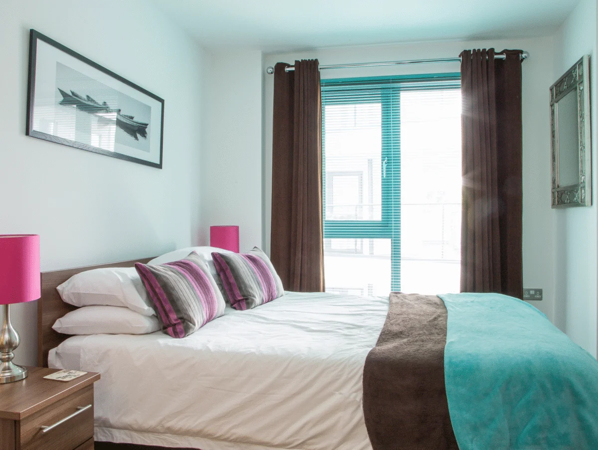 Bright double bedroom with pink cushions, artwork, and modern window at The Willow Suite, Cardinal Place, Woking