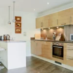 Contemporary open-plan kitchen with wood cabinetry, integrated appliances, and breakfast bar at The Willow Suite, Cardinal Place, Woking