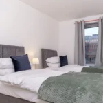 Twin bedroom with grey headboards, green throw blankets, and window at The Sycamore Suite, Enterprise Place, Woking