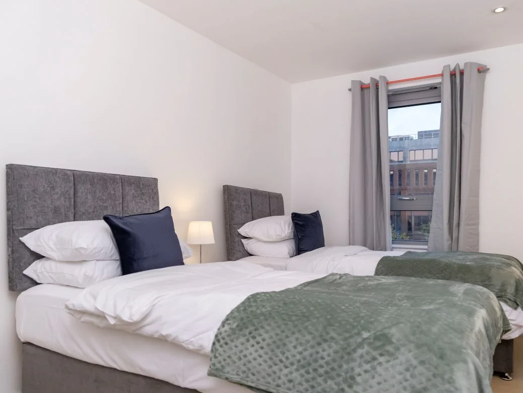 Twin bedroom with grey headboards, green throw blankets, and window at The Sycamore Suite, Enterprise Place, Woking